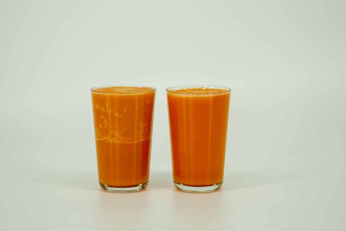 Two glasses of carrot smoothie twenty minutes after blending. The air-blended glass on the left has separated into a pale foam cap over settled pulp. The VOID glass on the right is one uniform color.
