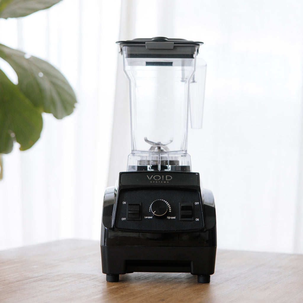 The VOID Complete System blender on a wooden counter with its basic lid on, a plant and a curtain behind it
