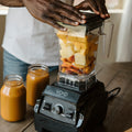 Hands resting on the lid of the VOID Complete System jar, filled with cut fruit, with two jars of finished smoothie on the counter