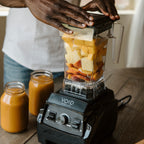 Hands resting on the lid of the VOID Complete System jar, filled with cut fruit, with two jars of finished smoothie on the counter
